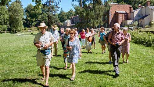 A group of senior visitors walking across a wide green field area with the red brick mill in the background